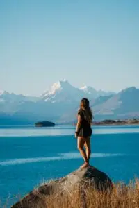A person stands barefoot on a rock, looking out over a blue lake towards snow-capped mountains
