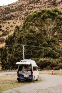 White camper van with rear hatch open, showing a bed and two camping chairs, parked on a dirt road against a scenic hillside