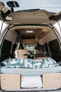 Interior view of a converted BANZ camper van featuring a bed with patterned pillows and wooden storage cabinets