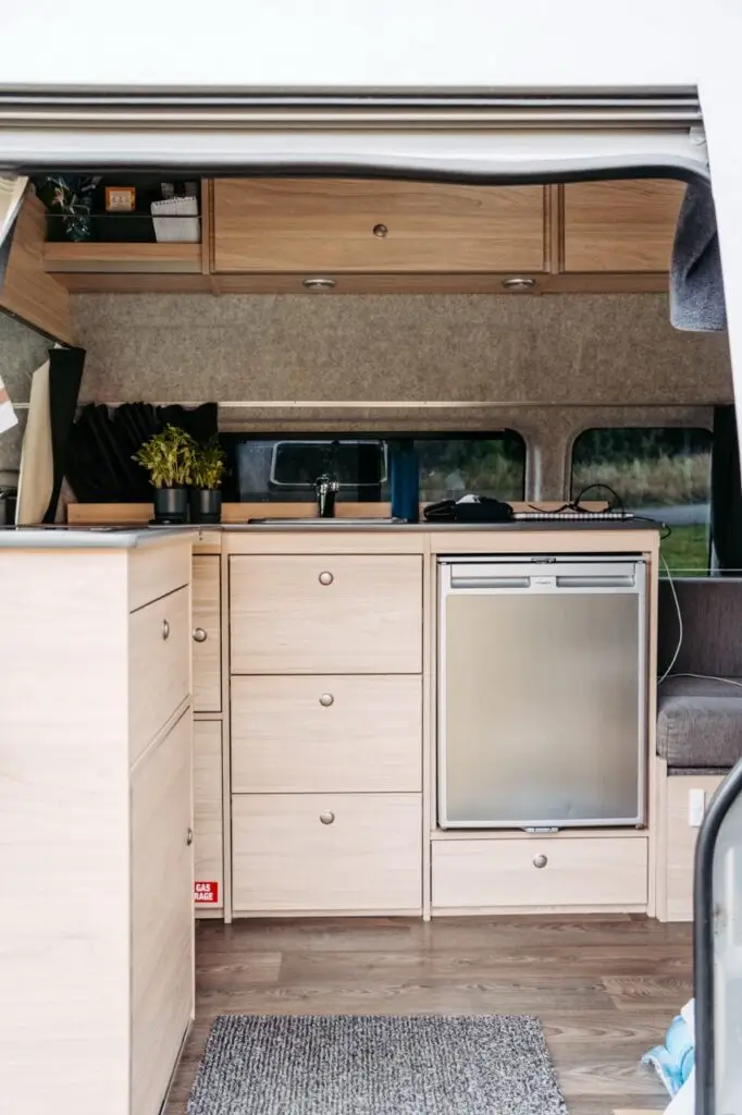 Interior of a BANZ New Zealand camper van featuring light wood cabinetry, a sink, and a small refrigerator
