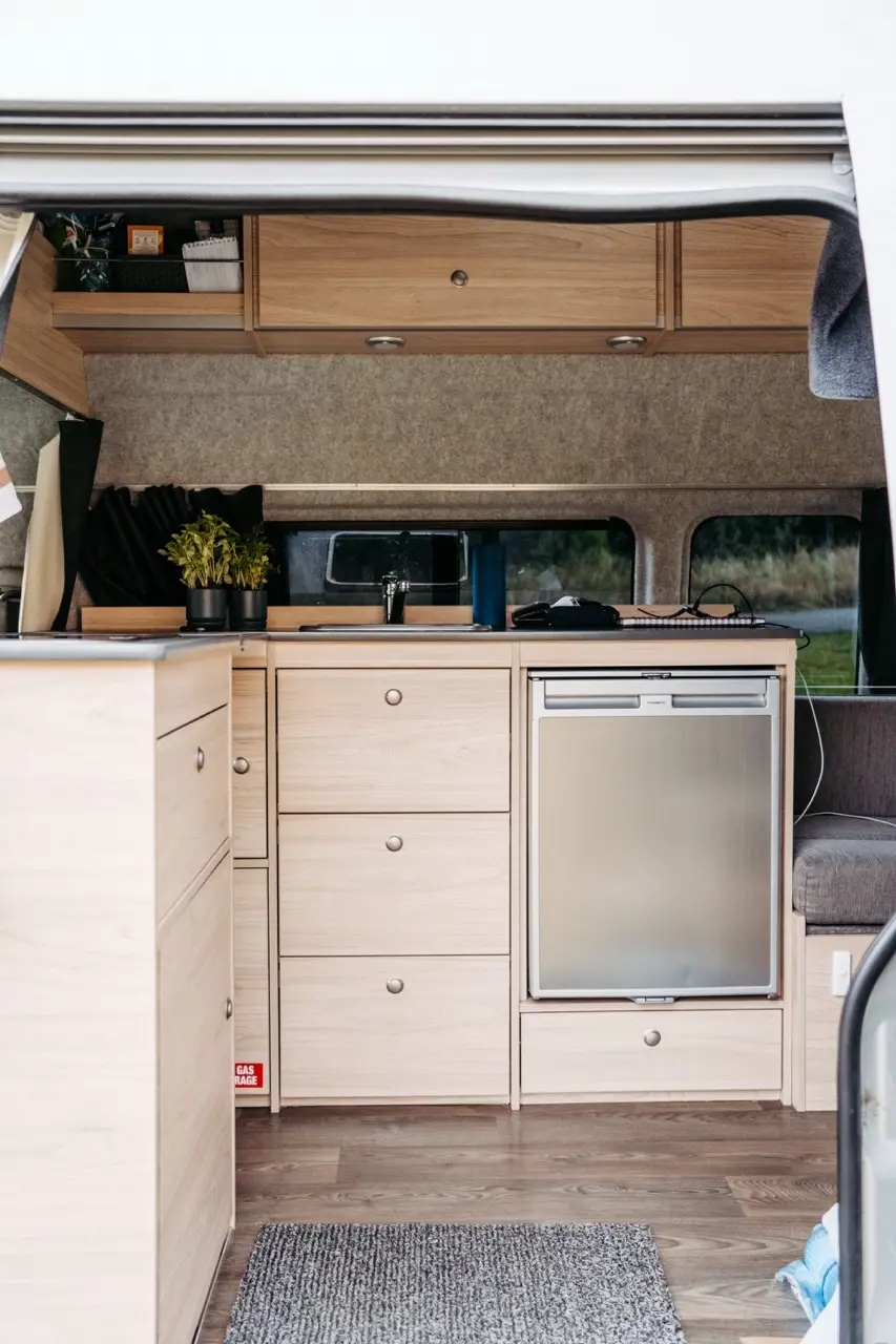 Interior of a BANZ New Zealand camper van featuring light wood cabinetry, a sink, and a small refrigerator