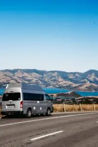 A silver Toyota Hiace campervan parked on a road overlooking a bay and rolling hills under a blue sky