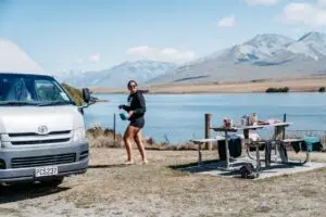 A woman walks barefoot with a bowl past a silver BANZ Travelcars campervan towards a picnic table by a lake with mountains