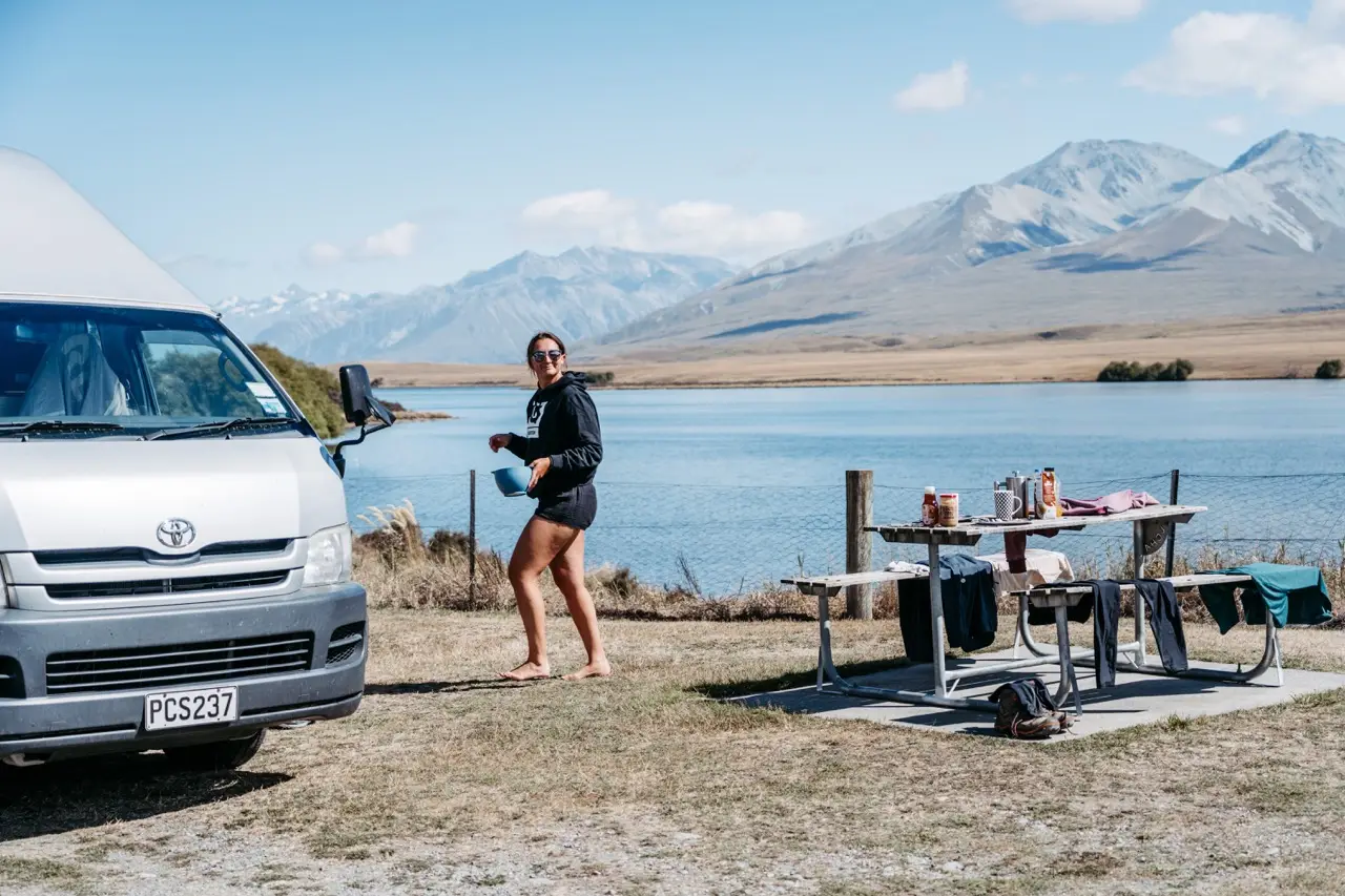 A woman walks barefoot with a bowl past a silver BANZ Travelcars campervan towards a picnic table by a lake with mountains