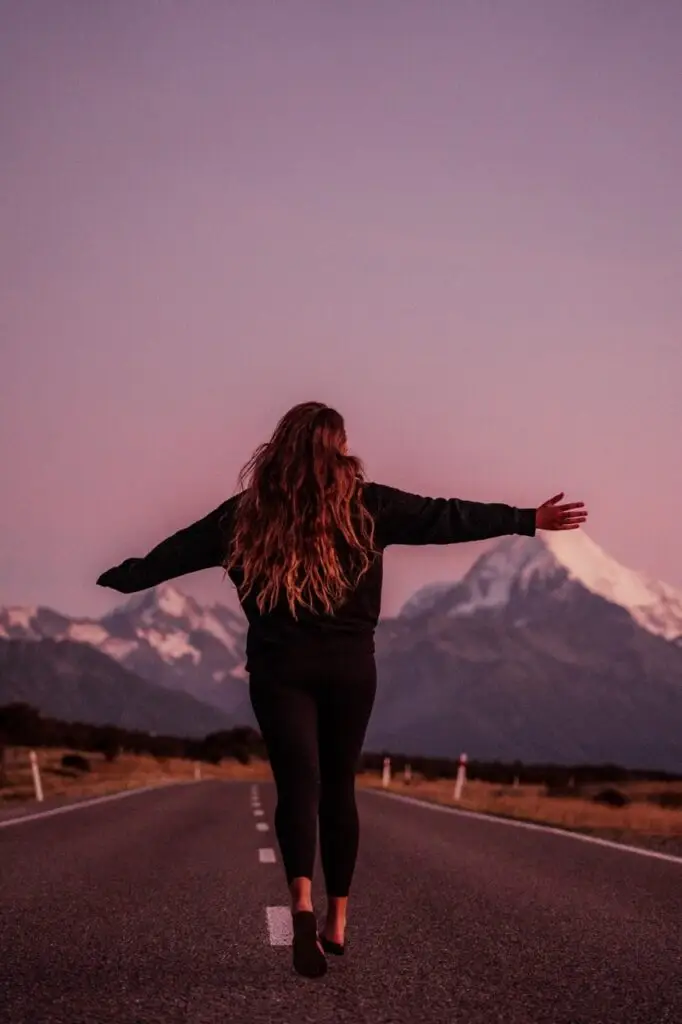 Person with long hair walking down an empty road with arms outstretched toward snow-capped mountains at dusk