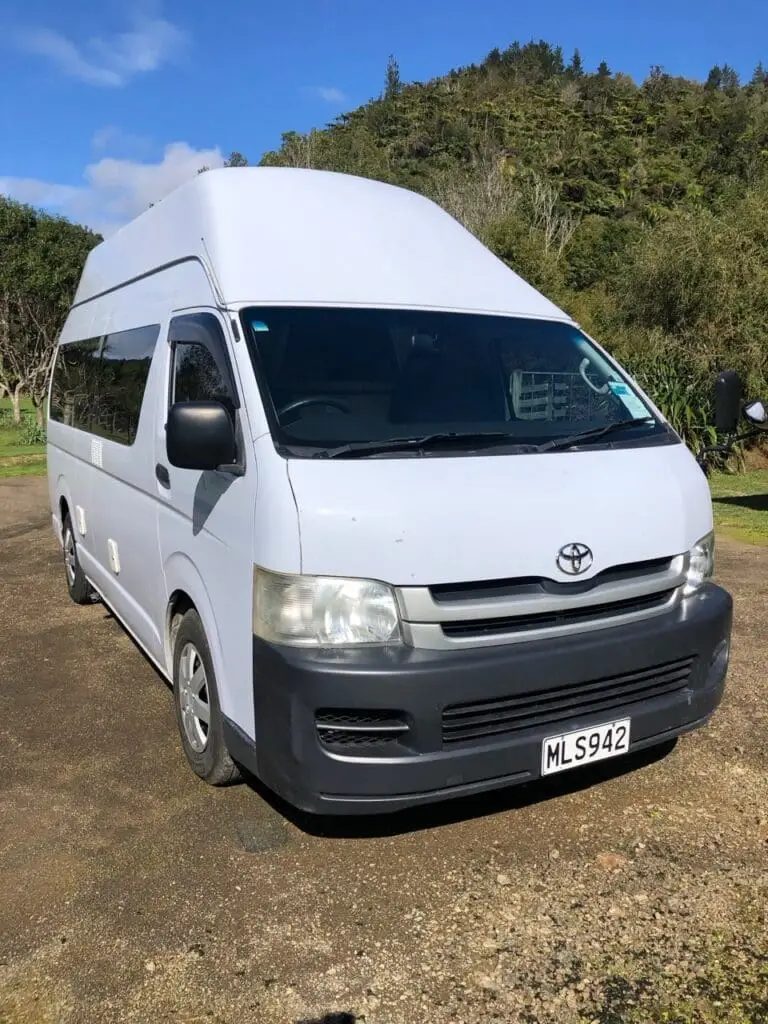 A white Toyota van with a high roof is parked on a sunny day on a gravel area, surrounded by greenery and trees, with a forested hillside in the background. The van’s license plate reads MLS942.