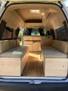 Interior of a camper van from BANZ Travelcars New Zealand with two cushioned bench seats, light wood paneling, overhead cabinets, and curtains. The back doors are open, revealing a cozy, well-lit living space.