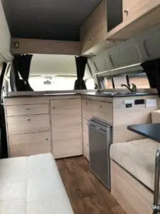 A compact camper van interior with light wood cabinets, a small fridge, countertop, and seating area. Dark curtains cover the windows and the floor has a wood-like finish, creating a cozy, modern atmosphere.