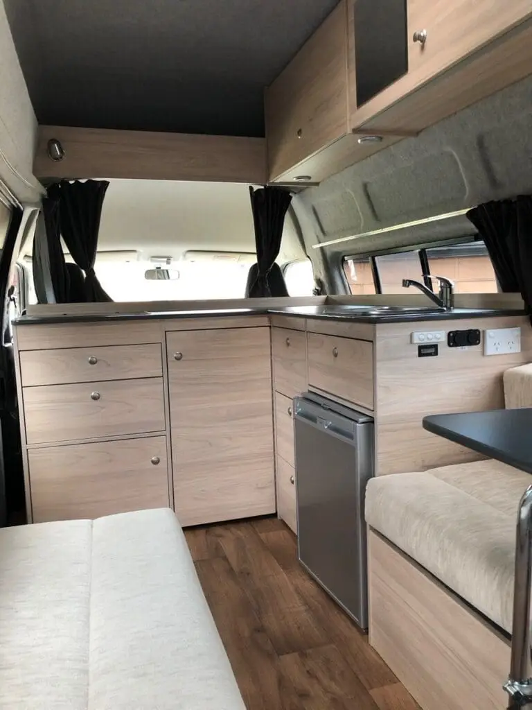 A compact camper van interior with light wood cabinets, a small fridge, countertop, and seating area. Dark curtains cover the windows and the floor has a wood-like finish, creating a cozy, modern atmosphere.
