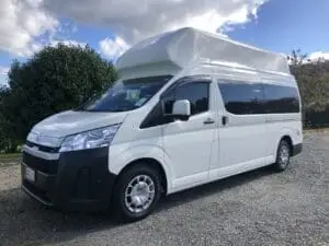 A white high-roof BANZ GRANDE campervan is parked on a gravel surface beside greenery under a partly cloudy sky, offering the perfect scene for those looking to rent a camper in New Zealand.