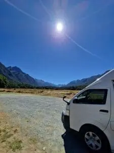 A bright sun shines over a scenic mountain landscape with a blue sky. In the foreground, part of a white van is visible, parked on a gravel area next to grassy fields and distant forested hills.