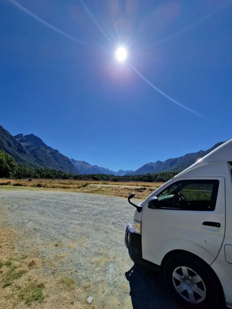 A bright sun shines over a scenic mountain landscape with a blue sky. In the foreground, part of a white van is visible, parked on a gravel area next to grassy fields and distant forested hills.