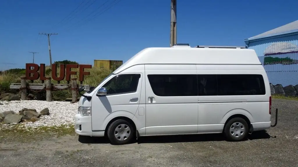 A white BANZ Travelcars New Zealand camper van is parked near large metal letters spelling BLUFF beside rocks and gravel, with a blue sky and some power lines in the background.