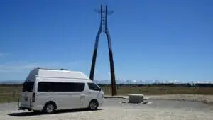 A BANZ Travelcars New Zealand campervan is parked on a gravel area near a tall, artistic metal sculpture with a face and wings, set against a backdrop of distant snow-capped mountains and a clear blue sky.