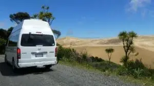A Camper van from BANZ New Zealand Campervan Hire is parked on a gravel road beside desert-like sand dunes and green vegetation under a bright blue sky with scattered clouds.