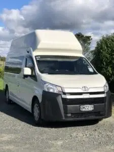 A white Toyota campervan with a high roof, from BANZ Travelcars New Zealand Campervan Hire, is parked on a gravel driveway beside greenery, under a partly cloudy sky.