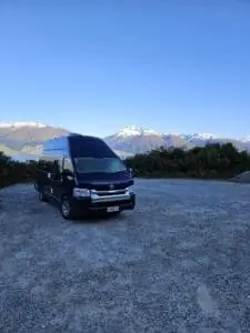 A dark blue van is parked on a gravel lot with snowy mountain peaks and green hills in the background under a clear blue sky.