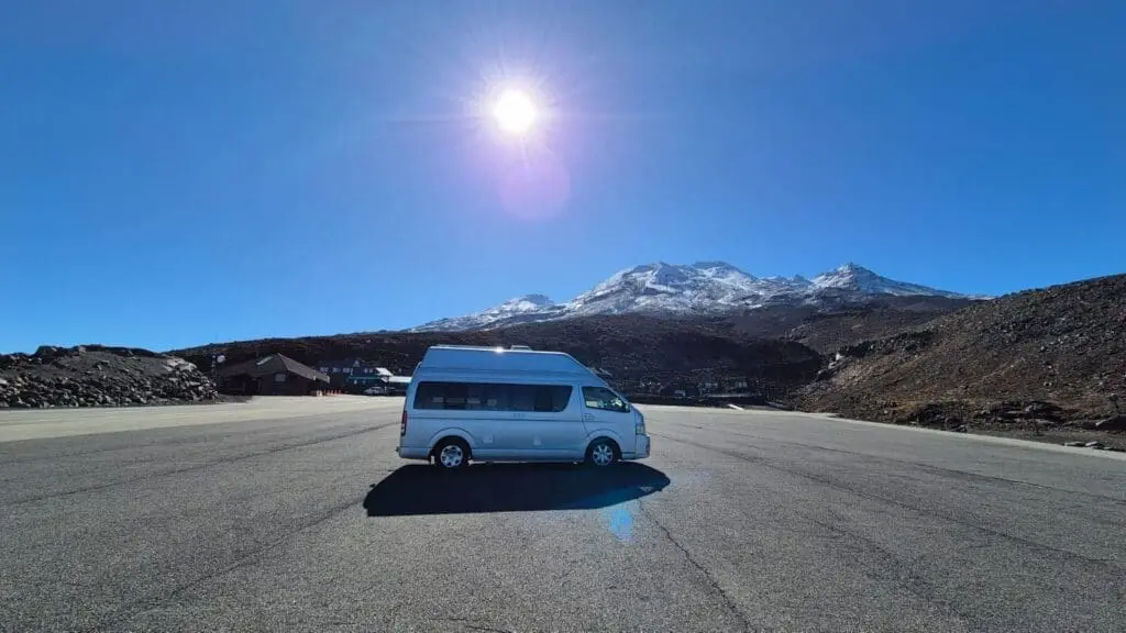 A white Banz Travelcars campervan parked on an empty paved area with snow-capped mountains in the background under a clear blue sky and bright sun.