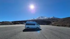 A white Banz Travelcars campervan parked on an empty paved area with snow-capped mountains in the background under a clear blue sky and bright sun.