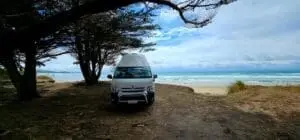 A white camper van, perfect for those looking to campervan mieten in Neuseeland, is parked under trees on a sandy patch by the beach, with waves rolling in under a cloudy blue sky in the background.