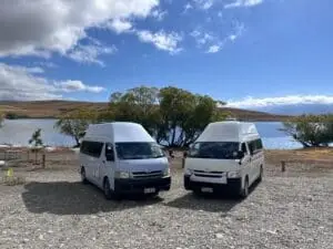 Two BANZ Travelcars campervans are parked on a rocky shore beside a lake, surrounded by trees and rolling hills under a partly cloudy sky—perfect inspiration to hire a campervan in the South Island of New Zealand for your own adventure.