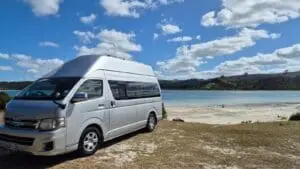 A silver campervan is parked on grassy ground near a calm, blue lake. In the background, there are green hills and a partly cloudy sky.