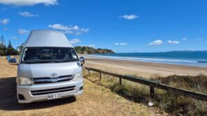 A silver BANZ Travelcars campervan is parked on dry grass near a sandy beach with gentle waves, lush green trees, and a bright blue sky with a few clouds in the background.