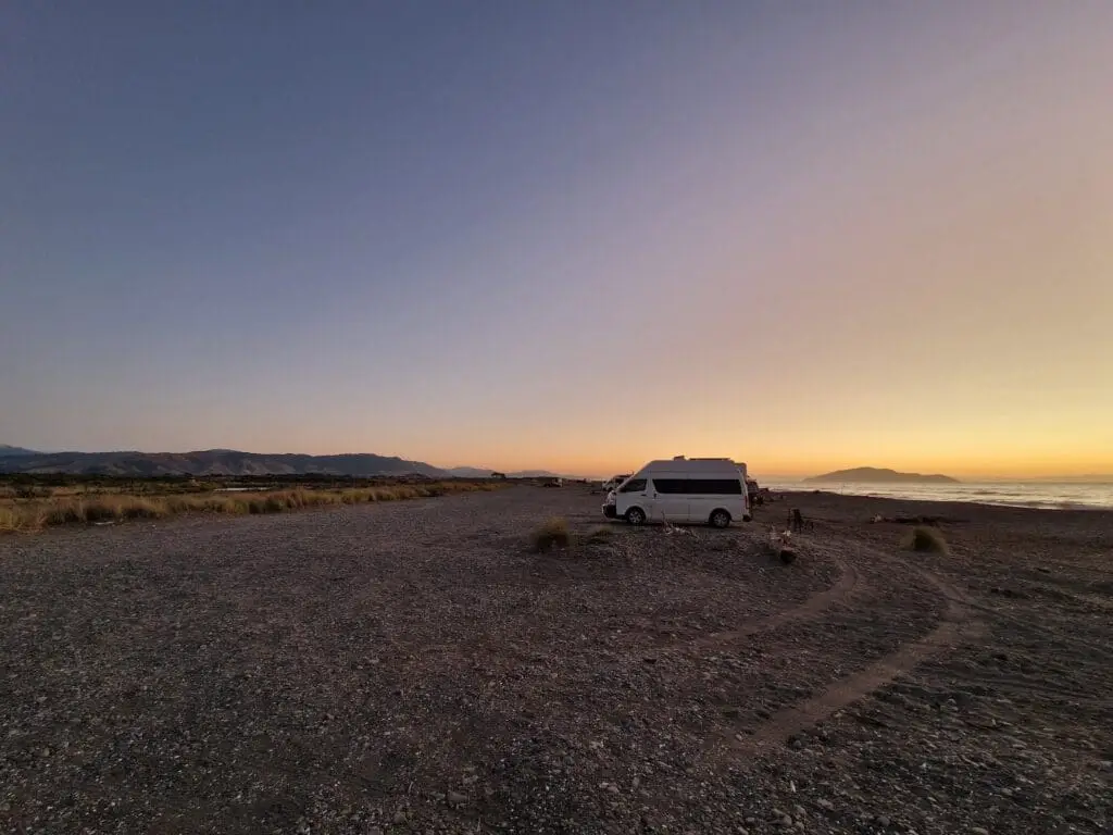 A camper van parked on a rocky beach at sunset, with mountains in the background, a golden sky, and calm ocean waves on the right side.