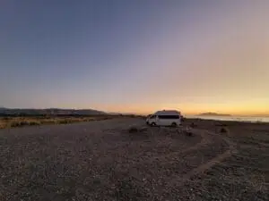 A camper van parked on a rocky beach at sunset, with mountains in the background, a golden sky, and calm ocean waves on the right side.