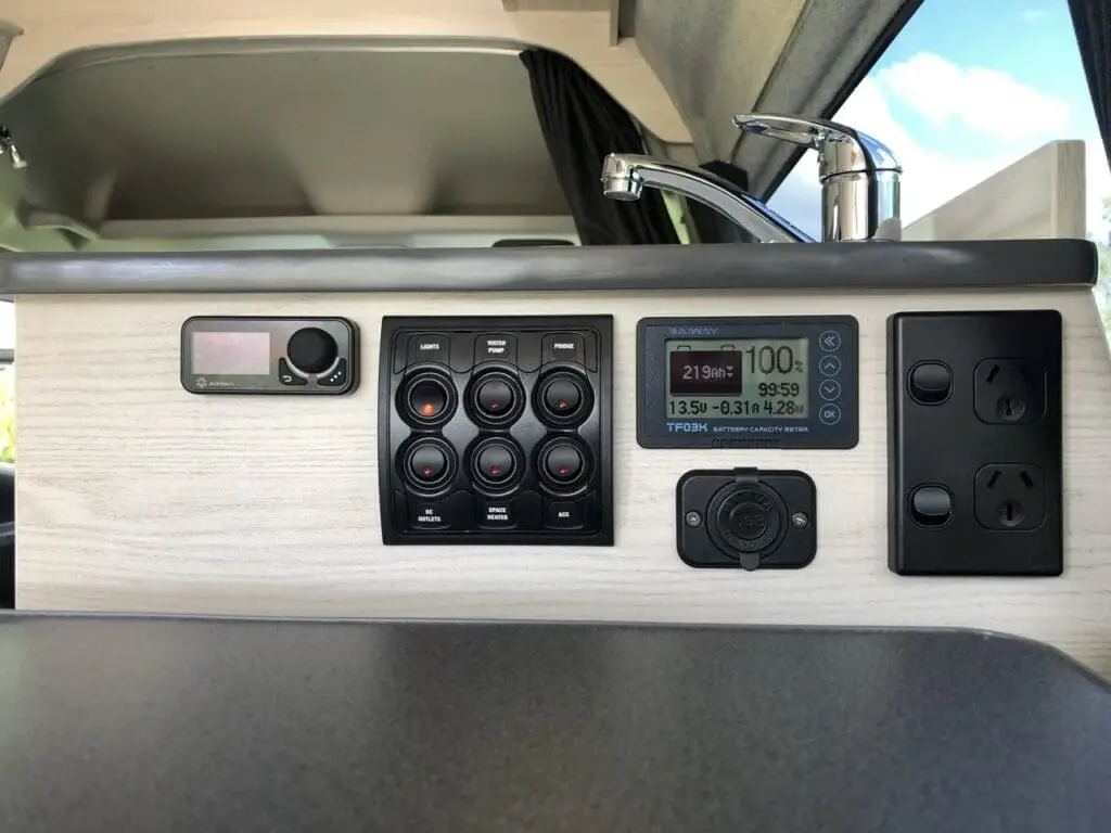 Close-up of a van’s interior control panel with switches, a digital display showing battery info, a faucet, power outlets, and part of a sink, all set against a light wood finish.