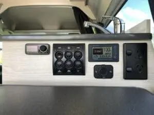 Close-up of a van’s interior control panel with switches, a digital display showing battery info, a faucet, power outlets, and part of a sink, all set against a light wood finish.