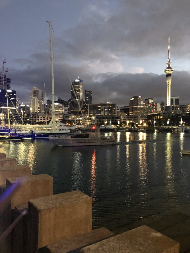 Auckland's Sky Tower and city skyline at dusk, with a marina full of boats and a moving boat reflecting lights on the water