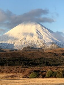 A snow-capped mountain peak bathed in sunlight under a blue sky with soft clouds, rising above dry brown hills and green trees