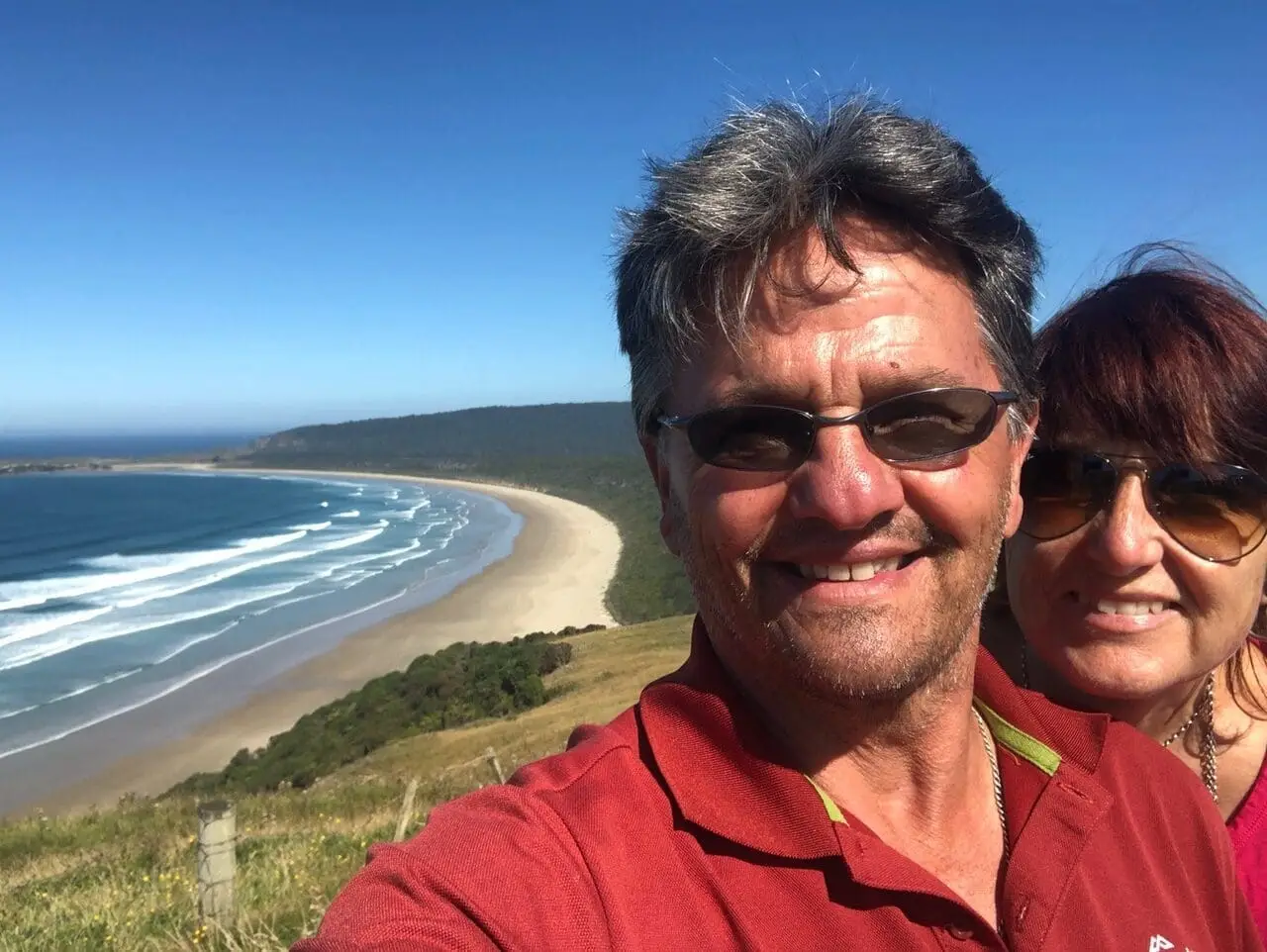 Kurt Binder and Yvonne Gloor smiling for a selfie on a grassy hill above a wide beach and ocean waves, enjoying the clear blue sky and distant greenery during their New Zealand Campervan Hire adventure.