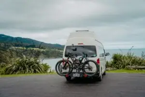A white BANZ Travelcars campervan with mountain bikes on a rear rack parked at a scenic coastal overlook