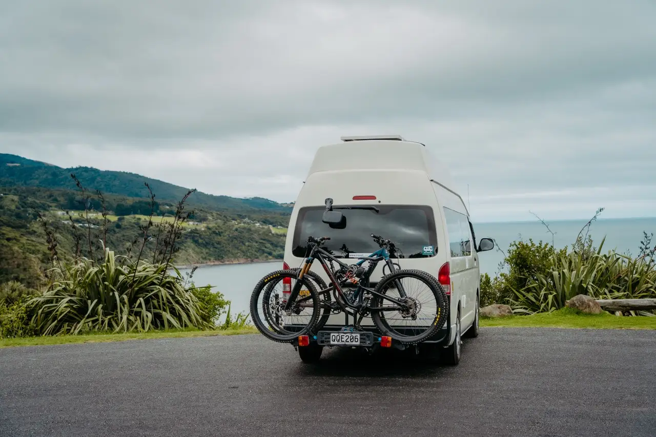 A white BANZ Travelcars campervan with mountain bikes on a rear rack parked at a scenic coastal overlook