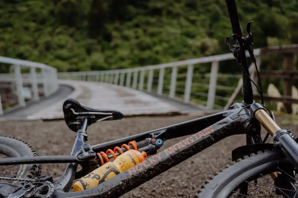A muddy Rocky Mountain bike with an orange suspension coil and yellow water bottle lies on a dirt path across a bridge