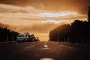A low-angle view of a road at sunset with parked cars on the left and trees lining both sides under an orange sky