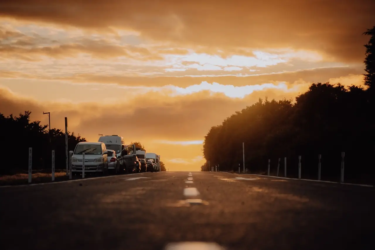 A low-angle view of a road at sunset with parked cars on the left and trees lining both sides under an orange sky