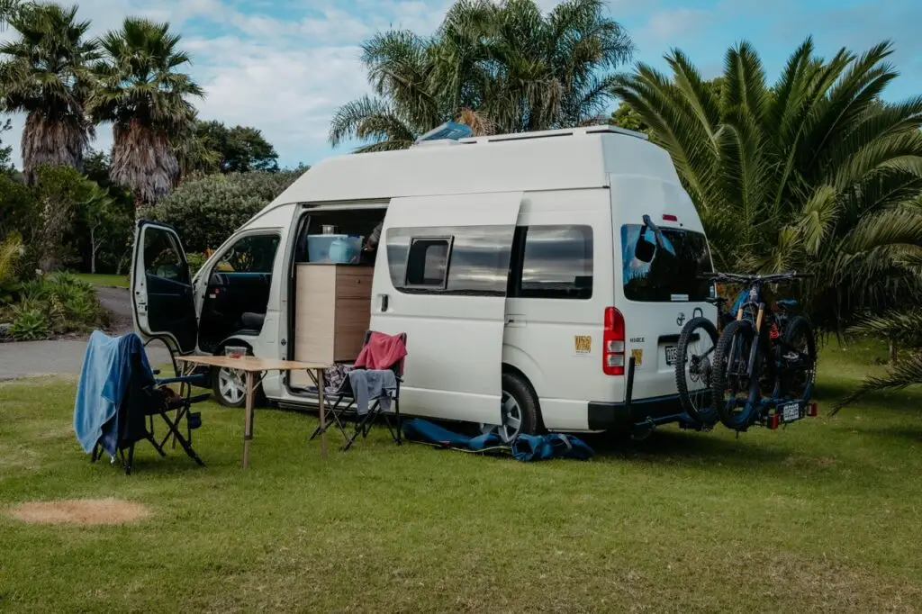 White camper van on green grass with open doors, two mountain bikes on its rear rack, and a table and chairs