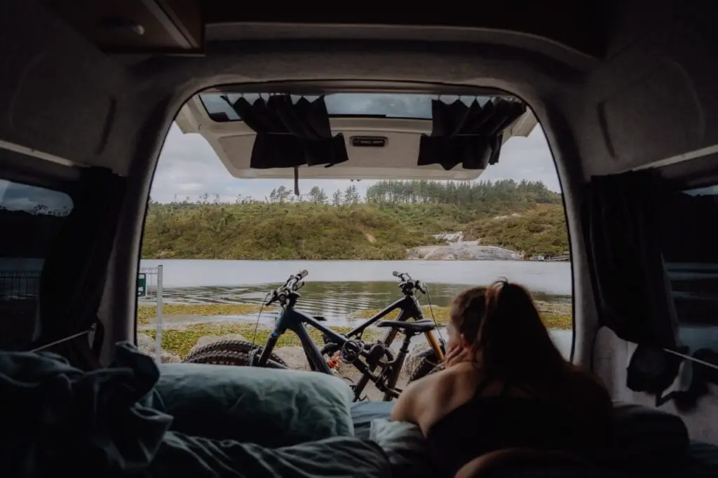A person lies in a camper van looking out at a lake with two mountain bikes and forested hills in the distance