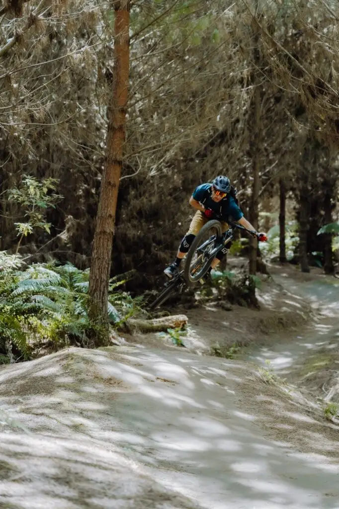 A mountain biker wearing a helmet and goggles jumps over a dirt mound on a wooded trail