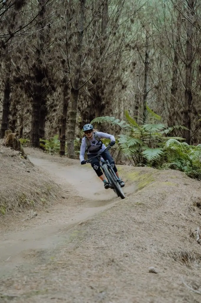 A mountain biker wearing a helmet and knee pads leans into a turn on a dirt trail in a forest