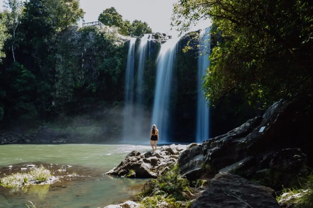 A woman stands on rocks looking at a multi-stream waterfall cascading into a green river surrounded by lush trees