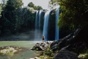 A woman stands on rocks looking at a multi-stream waterfall cascading into a green river surrounded by lush trees