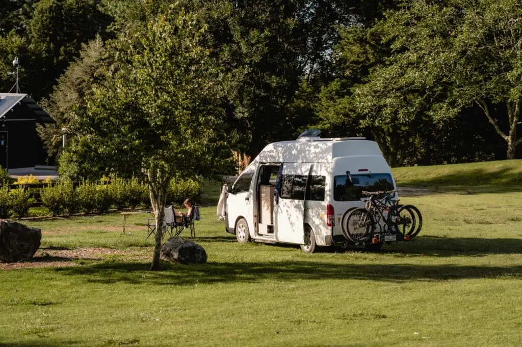 A white camper van with two bicycles on the back is parked in a grassy field, with a person relaxing under a nearby tree
