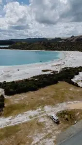 An aerial view of a turquoise lake with a white sand beach full of people, green hills, and a campervan parked nearby