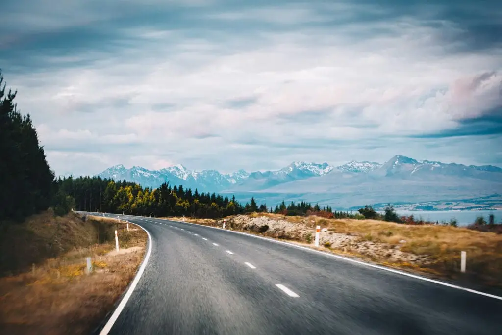 A winding asphalt road curves through grassy fields and forests, leading toward snow-capped mountains under a cloudy sky—perfect for exploring with a self contained campervan New Zealand rental.
