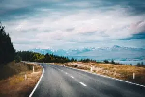 A winding asphalt road curves through grassy fields and forests, leading toward snow-capped mountains under a cloudy sky—perfect for exploring with a self contained campervan New Zealand rental.